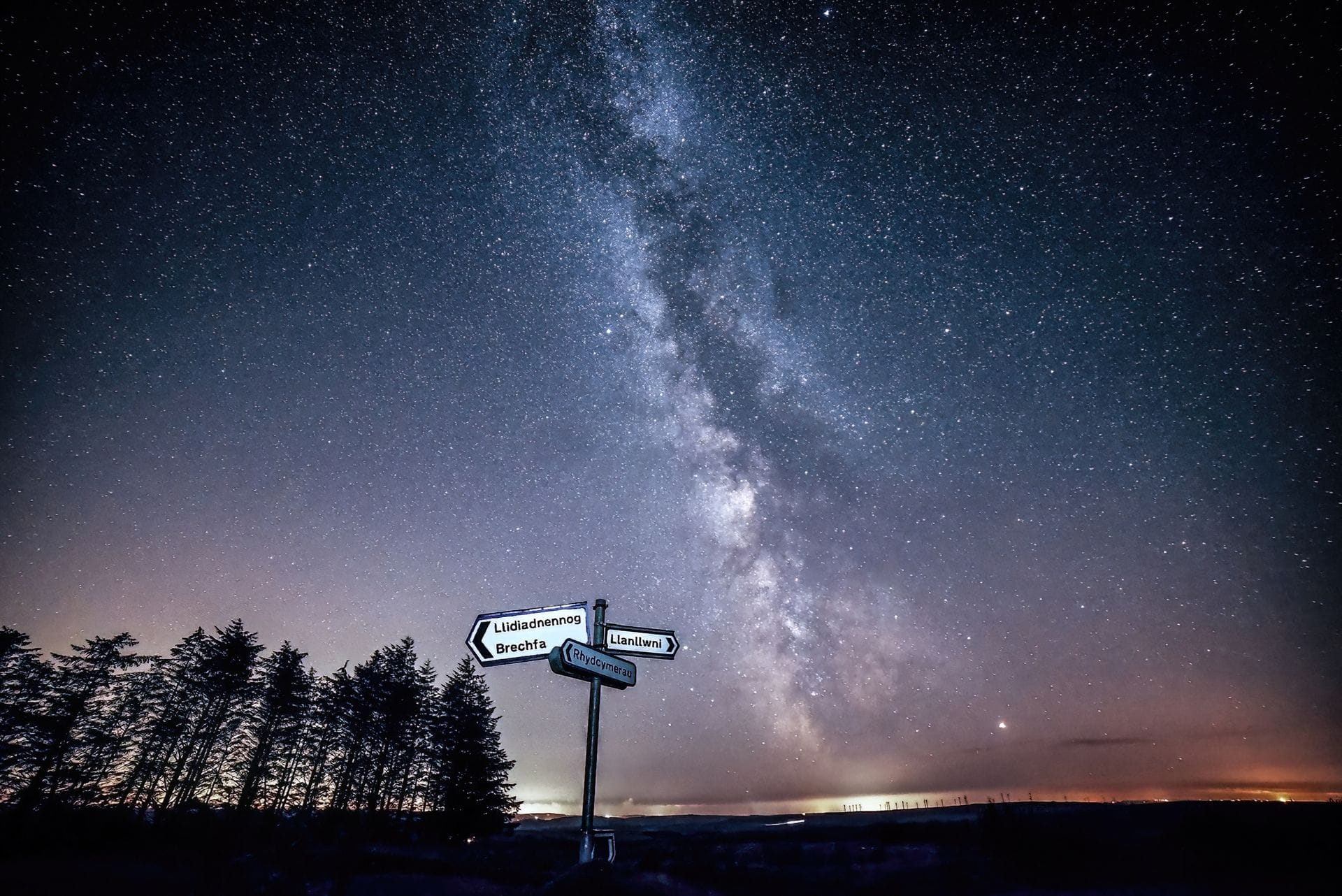A wide-angle view of star trails curving across the night sky above the open moorland of Bannau Brycheiniog National Park, with the silhouette of a single hawthorn tree in the foreground and the curve of a reservoir reflecting the stars below.