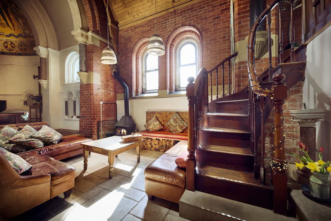 The interior of a converted Welsh chapel showing a spacious living area with exposed red brick walls, stone flagged floors, arched windows, and a prominent curved wooden staircase. The room is furnished with brown leather sofas, a wooden coffee table, and a large wood-burning stove.