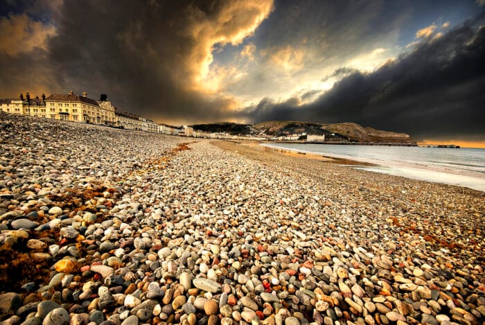 A dramatic sky with dark clouds looms over Llandudno beach, its wide pebbled shore lined with historic buildings on the left and distant hills in the background, whilst the calm sea stretches to the right.
