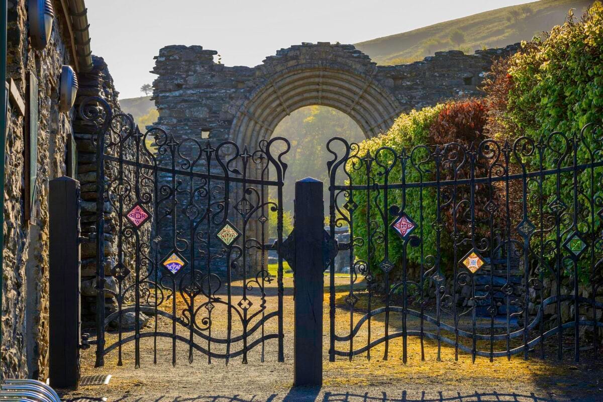 Ornate wrought iron gates with colourful tile inserts stand in the foreground, framing the historic Romanesque stone archway of the Strata Florida Abbey ruins in Ceredigion, Mid Wales, with sunlight hitting the soft green hills in the distance.
