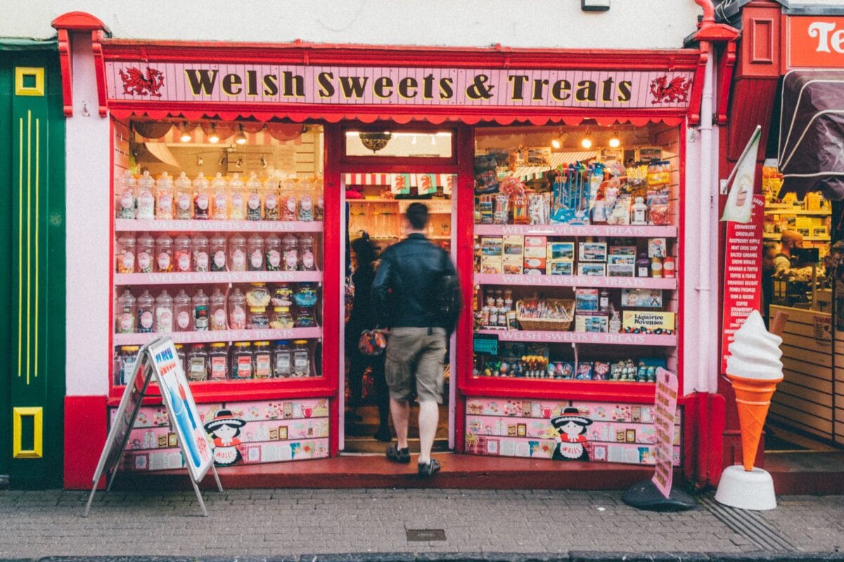 A person walking into the bright pink and red Welsh Sweets & Treats shop. The window displays are packed with rows of large glass sweet jars, local fudge boxes, and a large plastic ice cream cone standing on the pavement outside.
