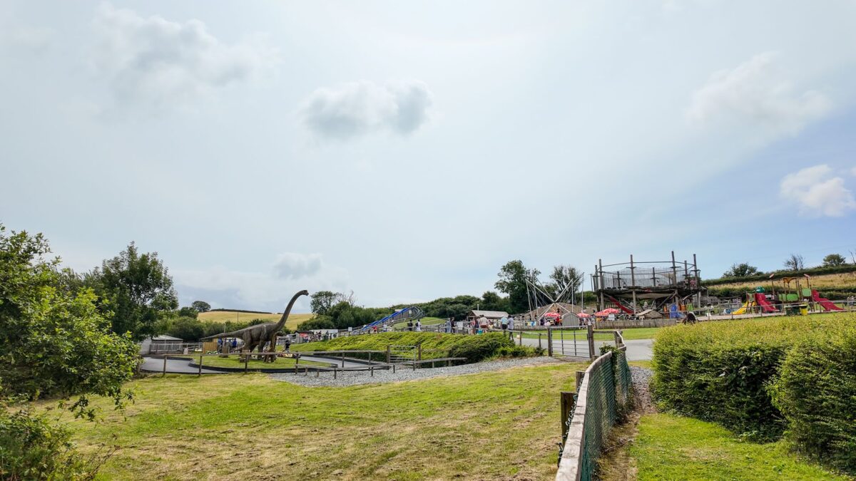A wide outdoor view of The Dinosaur Park in Tenby on a bright day. A tall, long-necked dinosaur model stands on the grass to the left, while visitors explore a large wooden climbing frame, slides, and outdoor play areas on the right.
