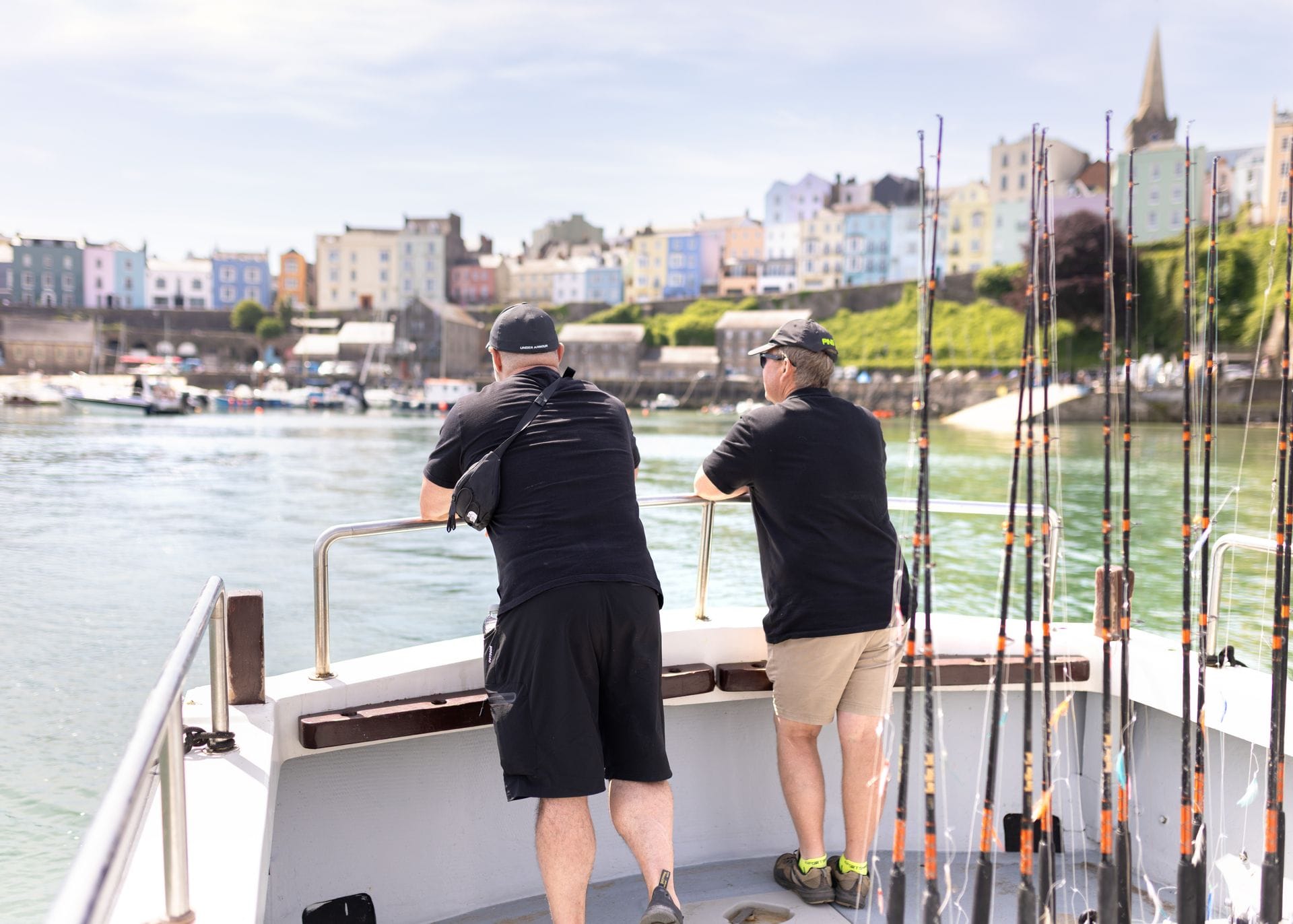 Two people leaning against the railing at the back of a charter boat equipped with tall fishing rods, looking toward the colourful, historic houses and church spire of Tenby from the sea.