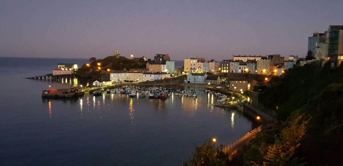Tenby Harbour at twilight, showing warm yellow lights from the colourful coastal buildings reflecting in the calm water where dozens of small fishing boats are moored.
