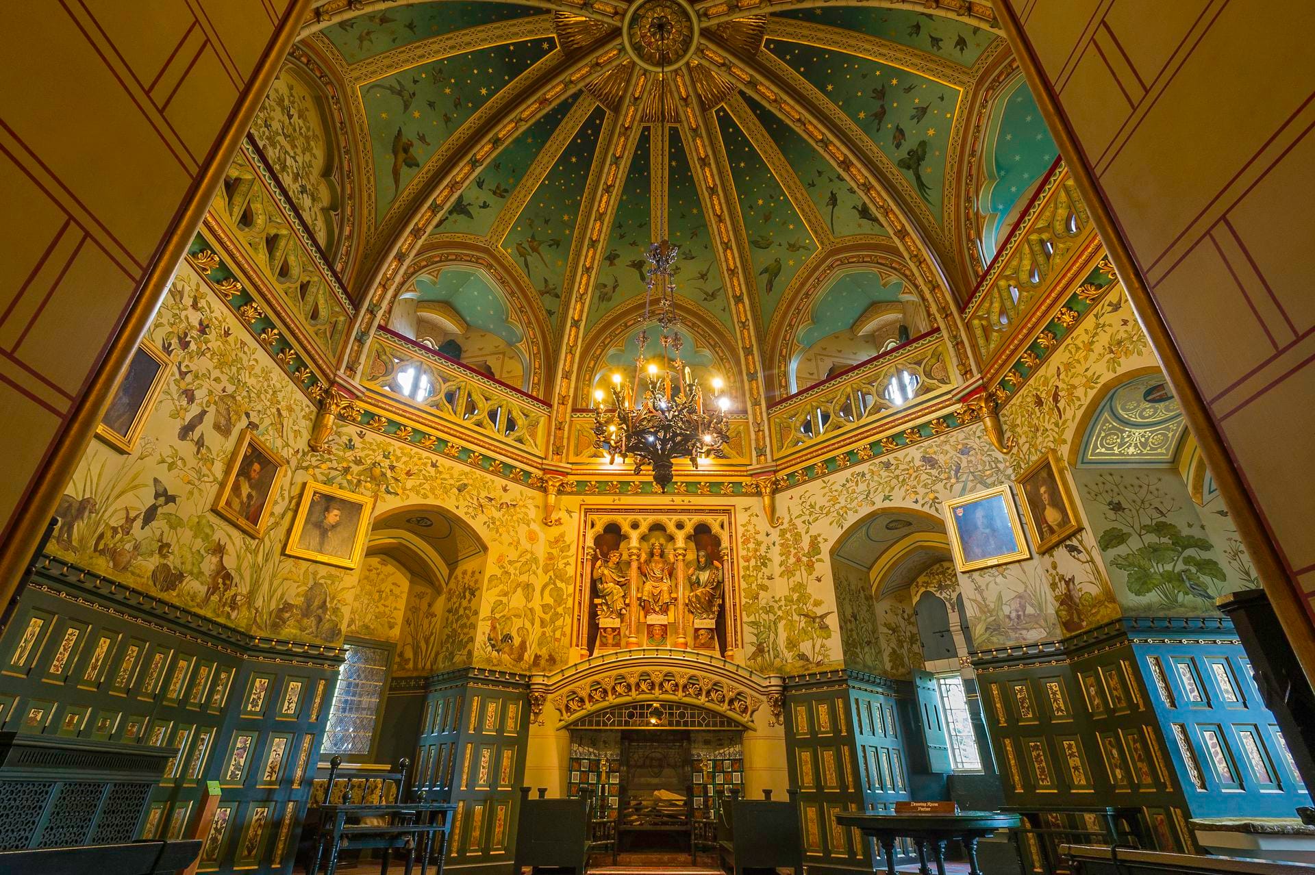 A wide view of the opulent Drawing Room inside Castell Coch. The highly ornate, octagonal room features a soaring vaulted ceiling painted with stars, elaborately decorated gold walls with murals, dark teal wood paneling, and a grand chandelier hanging in the center. A stunning setting for a Welsh wedding