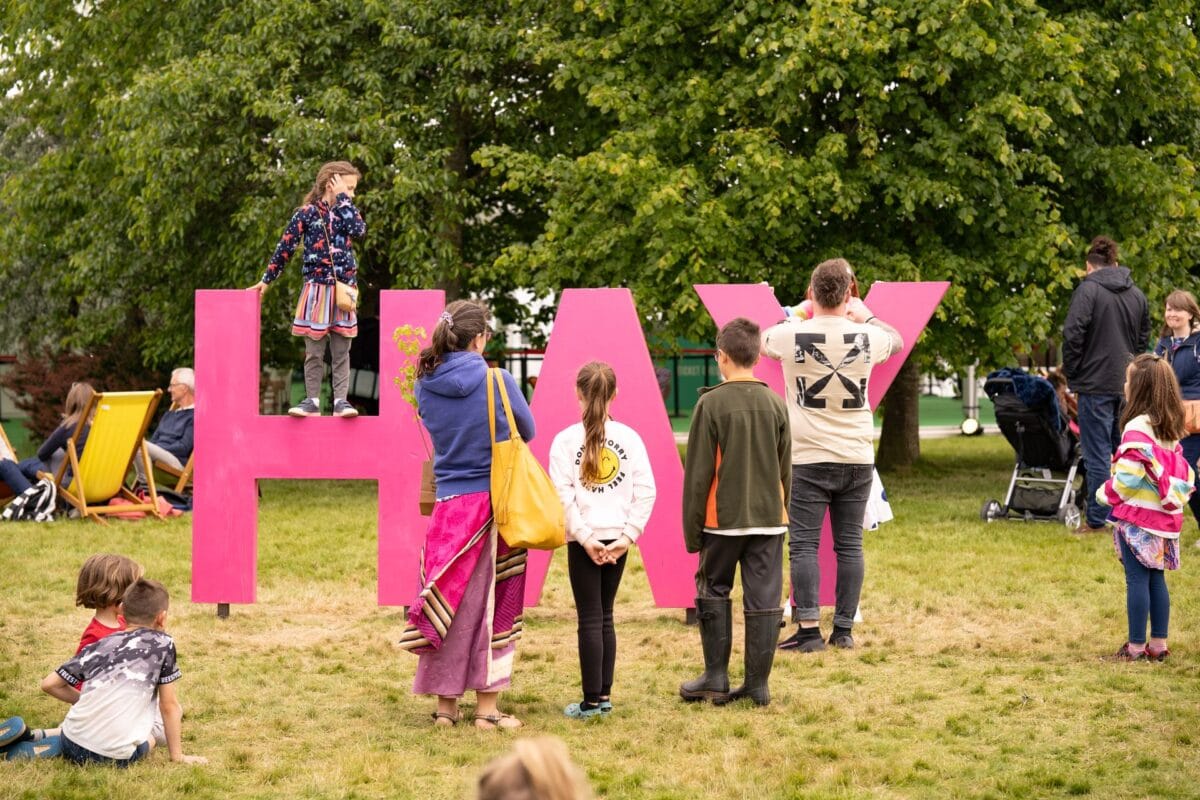 A group of adults and children in a park setting, standing and sitting on the grass around giant, bright pink letters spelling the word 'HAY'. One child is standing on the crossbar of the letter 'H'
