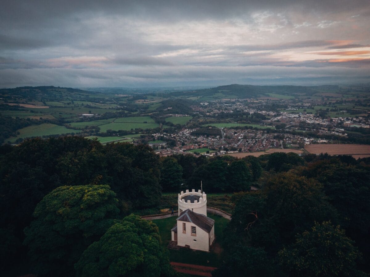 An elevated view of the white, castellated Round House at The Kymin, surrounded by dark woodland and overlooking the town of Monmouth and rolling green countryside beneath a cloudy sky.