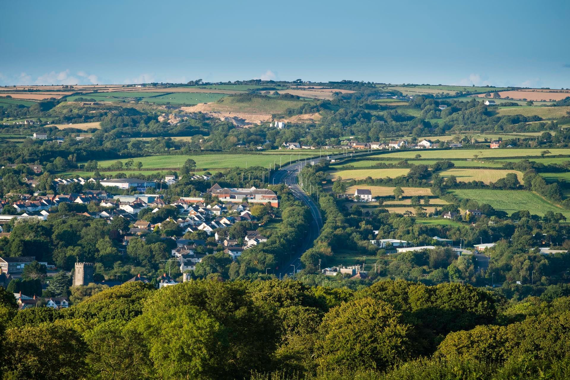 A scenic, elevated view overlooking the town of Cardigan in Wales, featuring lush green trees in the foreground, buildings and roads nestled in the valley, and rolling hills of patchwork farmland extending towards the horizon under a clear blue sky.