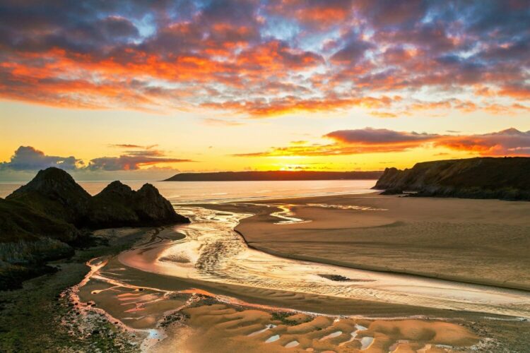 A breathtaking sunset over Three Cliffs Bay on the Gower Peninsula. A shallow stream meanders through a vast expanse of smooth sand toward the calm ocean. The bay is framed by the distinctive, jagged rock peaks on the left and steep dark cliffs on the right, all illuminated by a dramatic, vibrant sky filled with glowing orange, yellow, and deep purple clouds.