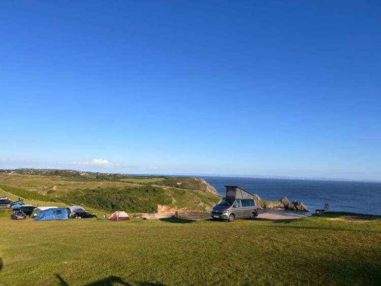 Several tents and a camper van are set up on a grassy hill overlooking cliffs and the blue sea, under a clear sky—near a beach, where sunlight casts long shadows along the scenic coastline.
