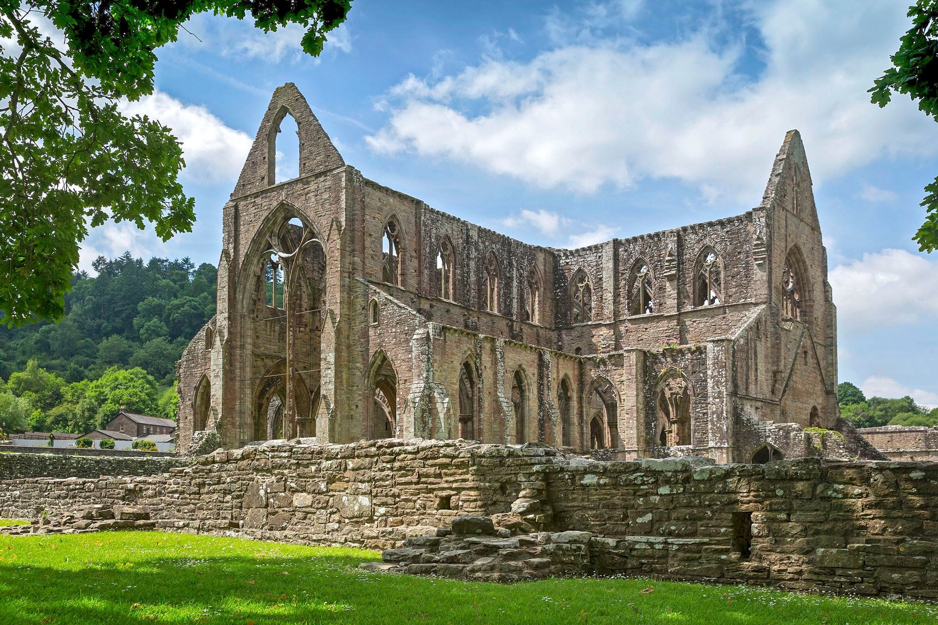 A spectacular view of the historic Tintern Abbey ruins, featuring grand roofless Gothic stone architecture and intricate window arches. The magnificent abbey is surrounded by vibrant green grass and a low stone wall, set against a backdrop of lush forested hills and a bright blue sky with fluffy white clouds.