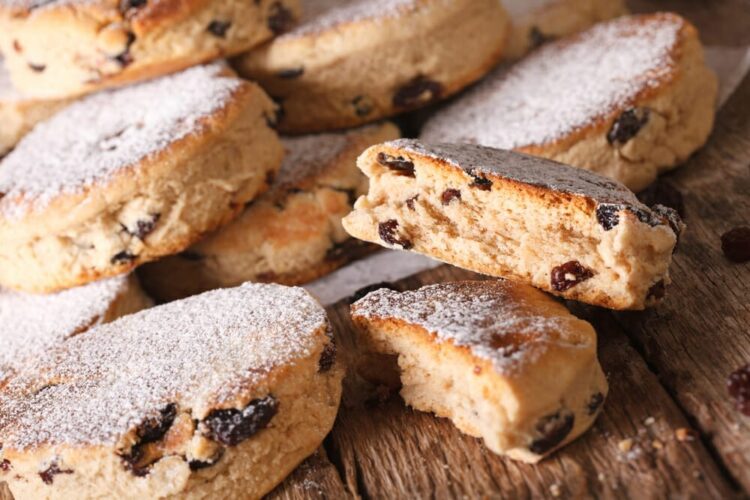 A close-up of several Welsh cakes with sultanas, dusted with icing sugar, arranged on a rustic wooden surface. One cake is broken in half, showing its soft, crumbly texture inside.