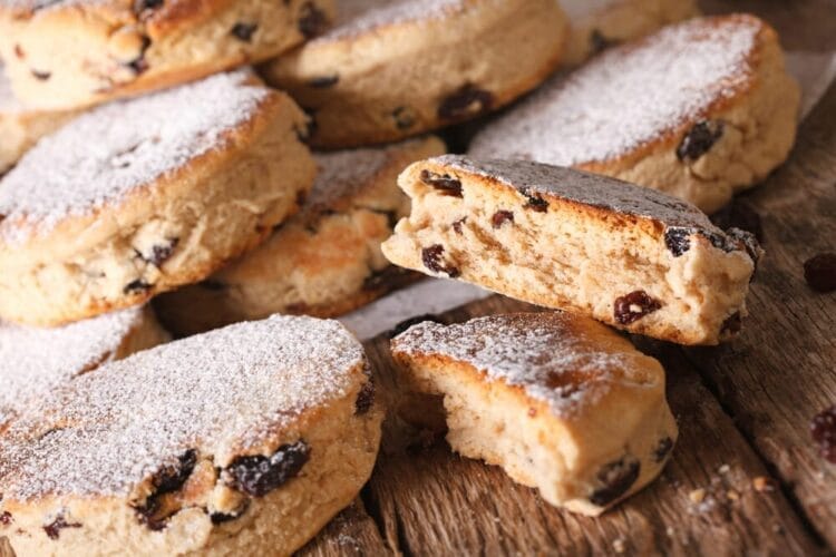A close-up of several Welsh cakes with sultanas, dusted with icing sugar, arranged on a rustic wooden surface, one cake broken in half showing its soft, crumbly texture