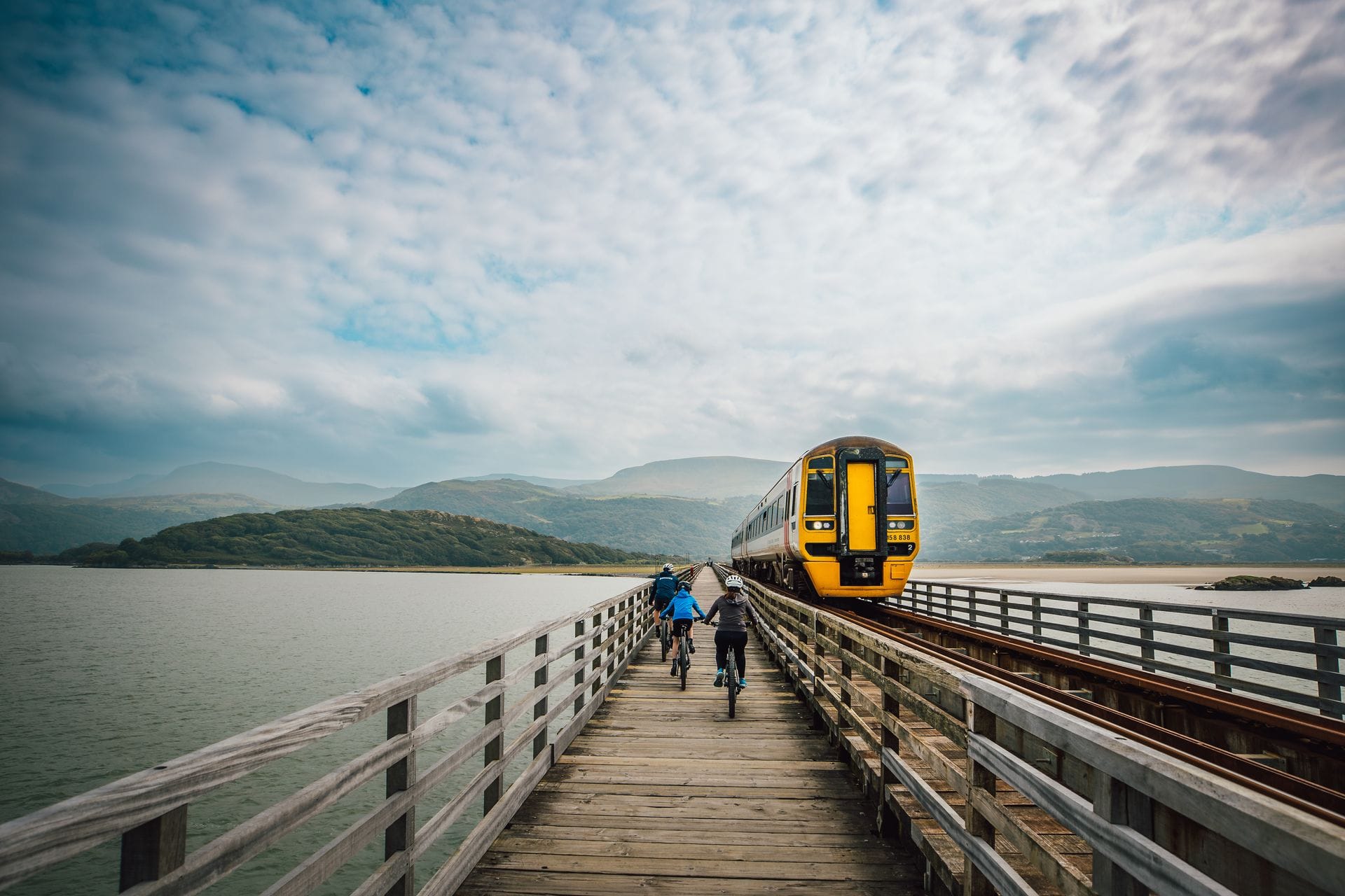 A yellow passenger train crosses the Barmouth Bridge on the right side of the frame, while a group of cyclists rides towards the camera on the adjacent wooden pedestrian path to the left. The bridge spans a wide estuary with rolling green hills and mountains in the background under a dramatic, cloudy sky.