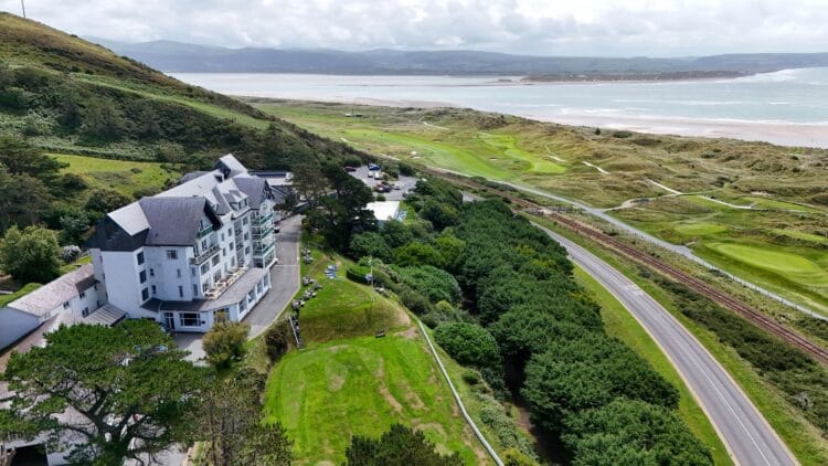 Trefeddian Hotel is known as one of the best hotels in Wales, sits on a hillside overlooking a golf course, winding road, lush greenery, and a sandy coastline with the sea and mountains in the background under a partly cloudy sky.