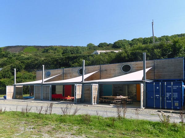 The exterior of Tŷ Cwch Boathouse on a sunny day, showing its unique construction from repurposed shipping containers. The upper level is clad in light wood with small round porthole windows, while the open ground floor features a seating area shaded by stretched white sail canopies. A blue shipping container anchors the right side, all set against a steep, green wooded hillside.
