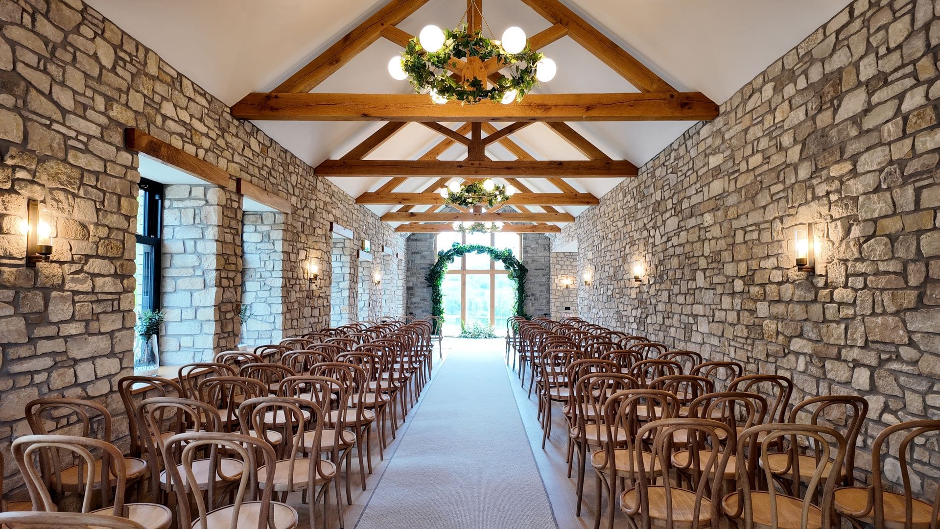 A long, rustic wedding ceremony room with exposed stone walls and a high vaulted ceiling featuring wooden beams and globe chandeliers decorated with foliage. Rows of wooden cross-back chairs flank a central white aisle that leads to a large arched window framed by a greenery arch.