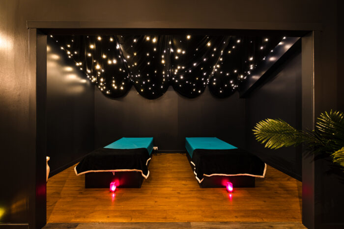 A dimly lit room at Vale Resort near Cardiff, with two massage tables covered in black and teal linen, a wooden floor, dark walls, a ceiling draped with fabric and twinkling lights, and a green plant in the corner.