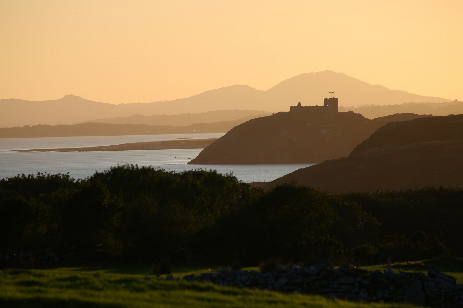 The silhouette of Criccieth Castle atop its headland at sunset, with layered mountains and Tremadog Bay fading into golden haze behind it.