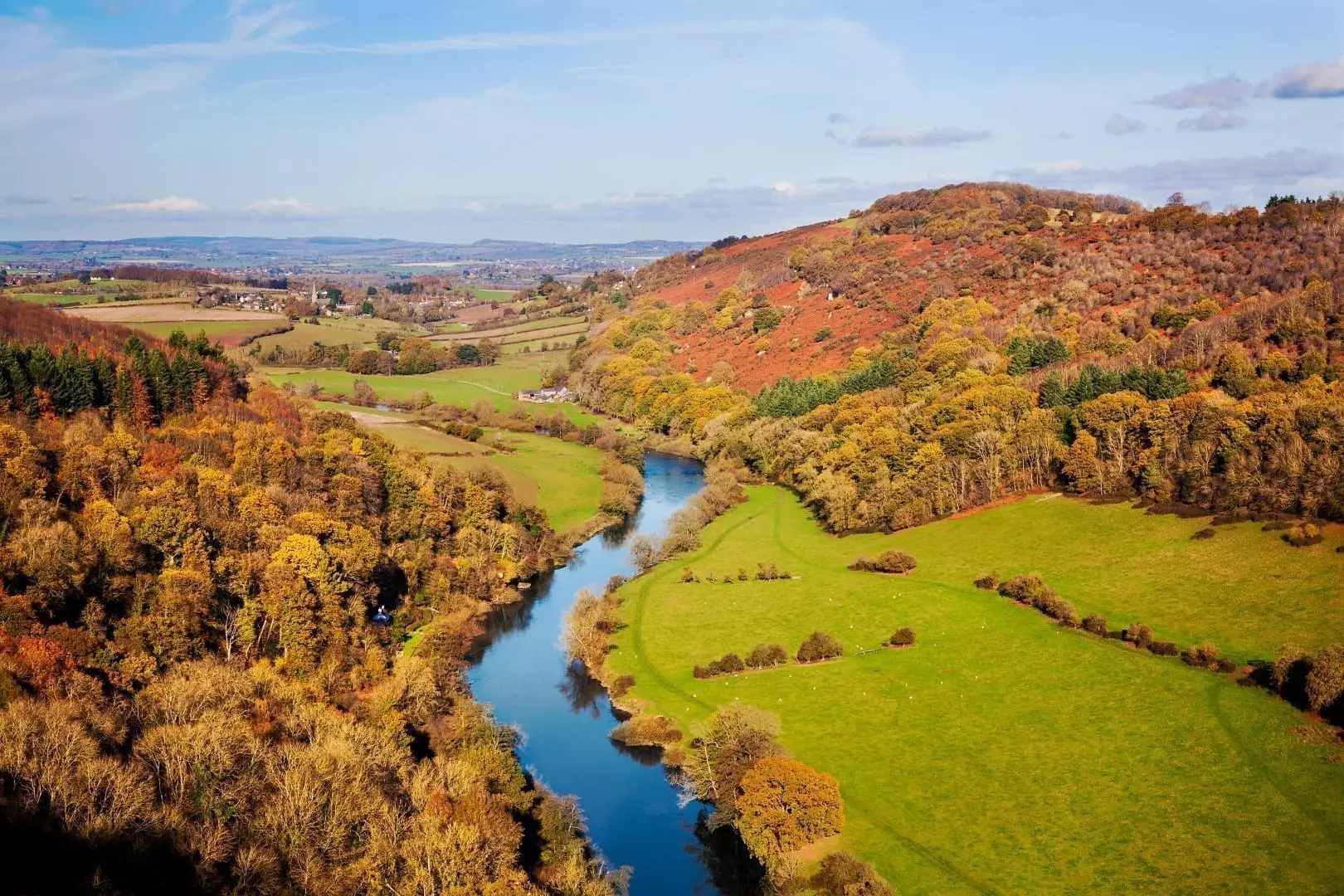 A scenic aerial view of the River Wye winding through bright green pastures and rolling hills covered in vibrant autumn woodlands under a clear blue sky, showcasing a beautiful landscape perfect for camping in the Wye Valley.