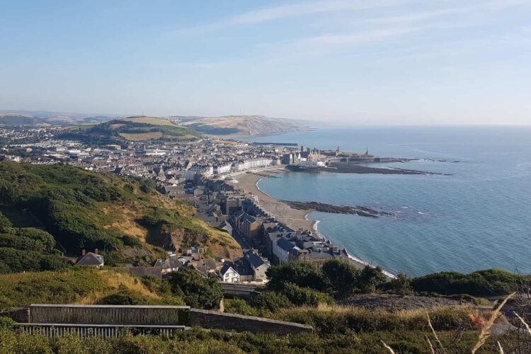 A coastal town, Aberystwyth features a sweeping sandy beach, a curved shoreline, and rows of houses, surrounded by green hills under a clear blue sky. The sea stretches out to the horizon.