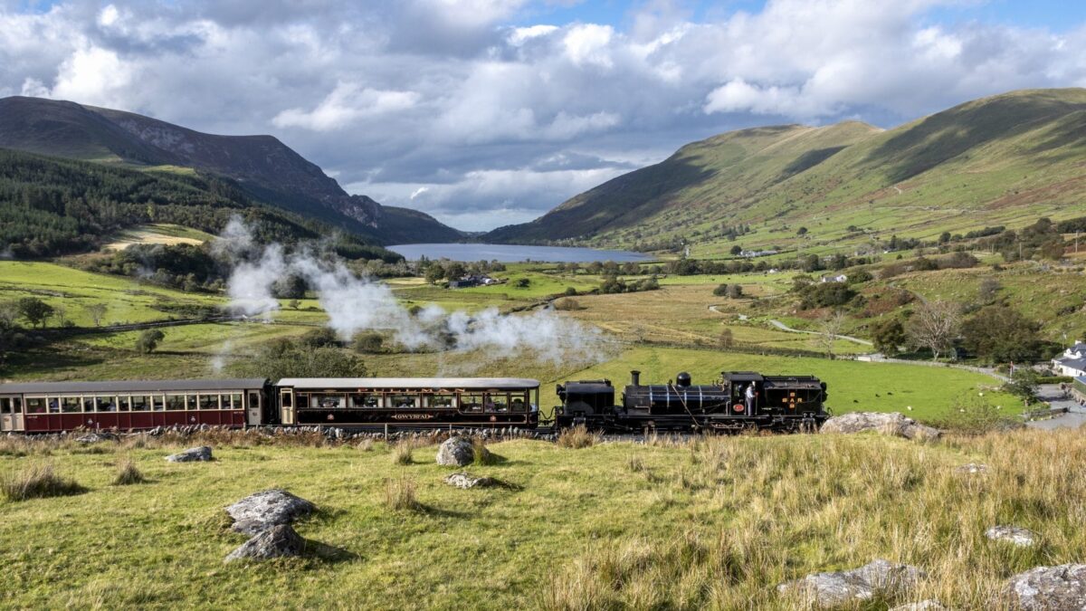 The historic Number 87 steam train on the Welsh Highland Railway puffing white smoke as it travels through a stunning green mountain valley, a scenic journey perfect for family days out in North Wales.