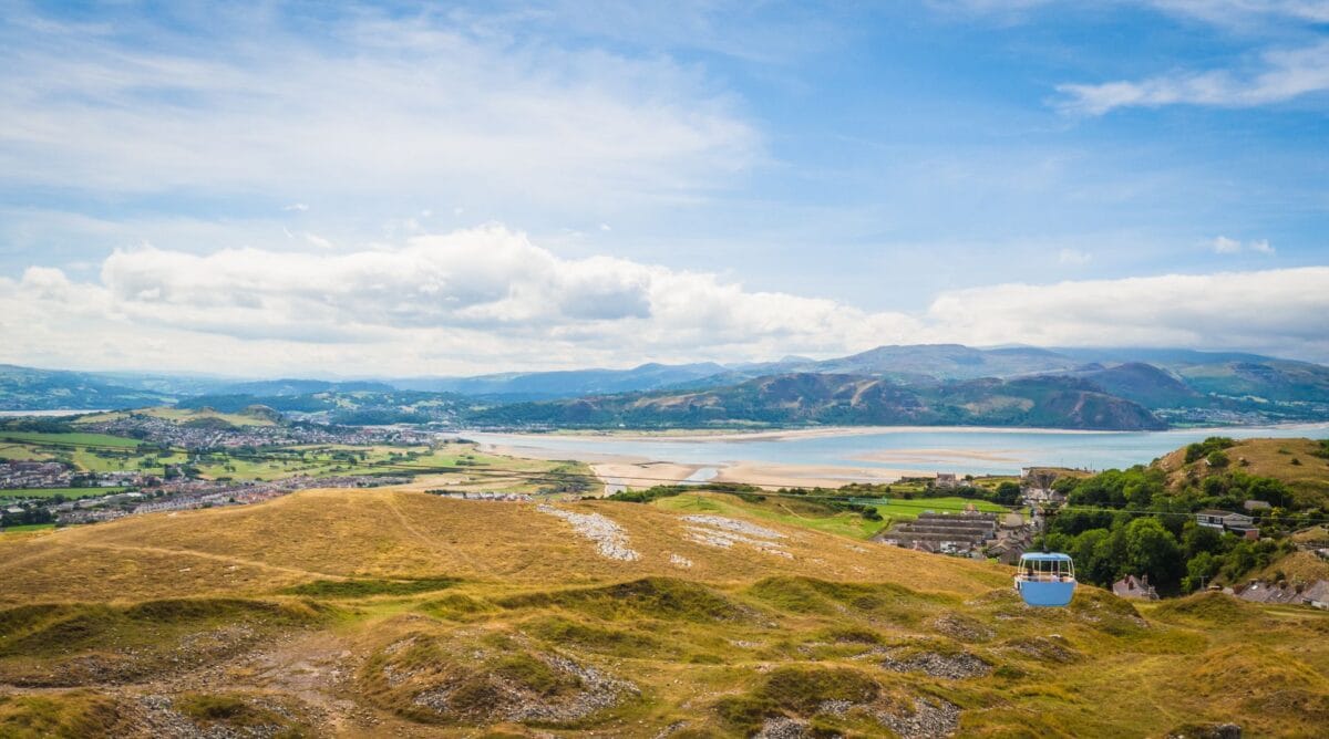 A light blue cable car traveling over grassy hills, with a scenic view of Llandudno, distant mountains, and a sandy estuary near the one of the best beaches in Wales.