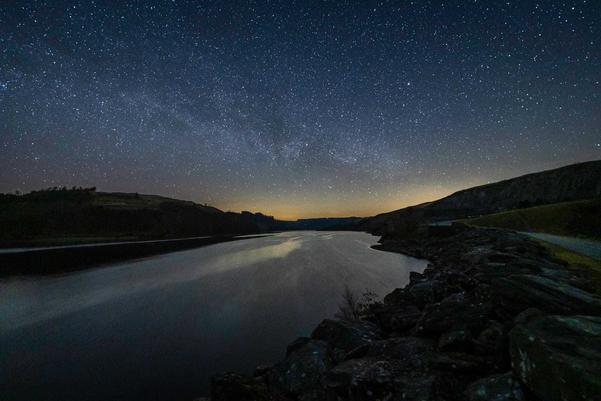 The reservoir at Elan Valley reflecting a star-filled night sky with the Milky Way visible across the horizon, surrounded by gentle hills and the silhouettes of conifer trees, captured in autumn during the new moon period.