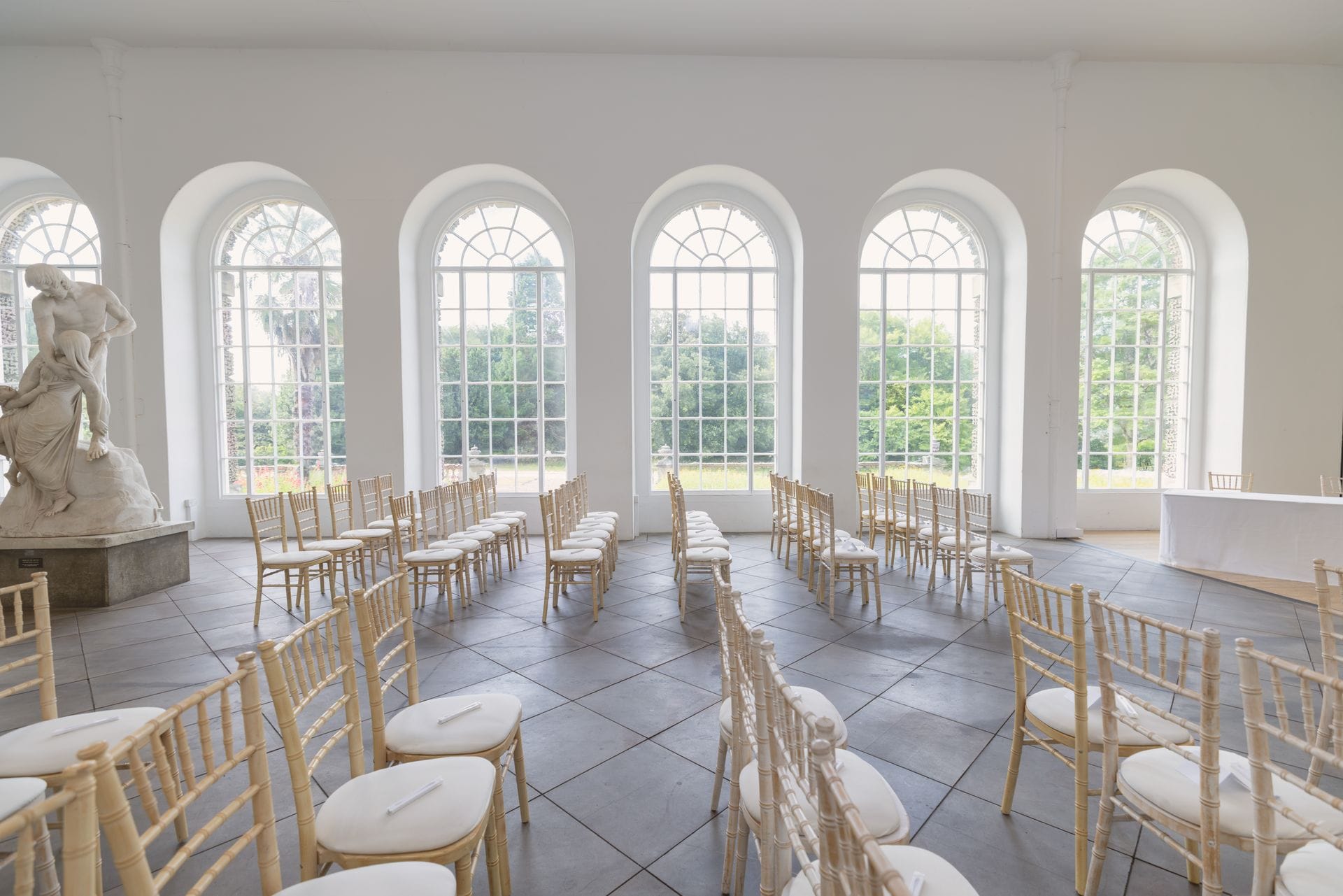 A bright, elegant room set up for a wedding ceremony. Light wooden Chiavari chairs are arranged in rows along a central aisle on a tiled floor. One wall is lined with large, floor-to-ceiling arched windows looking out onto lush green gardens, and a classical white statue stands on a plinth to the left.