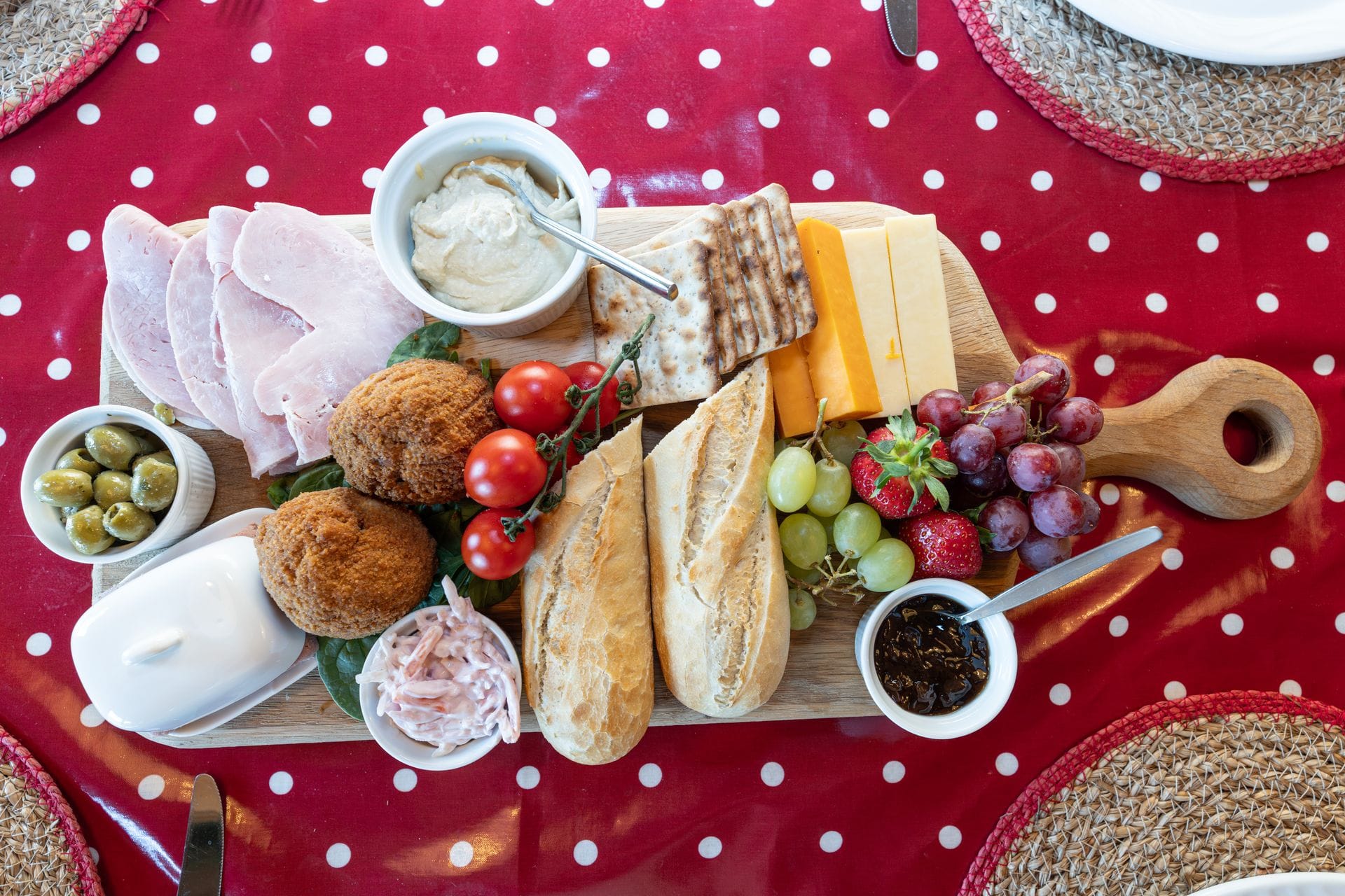 An overhead view of a wooden grazing board on a red polka-dot tablecloth, featuring sliced cold meats, a bowl of soft whipped cheese, cubes of orange and pale cheddar-style cheese, breaded cheese or croquette balls, a sliced baguette, cherry tomatoes on the vine, olives, grapes, strawberries and a bowl of dark chutney.
