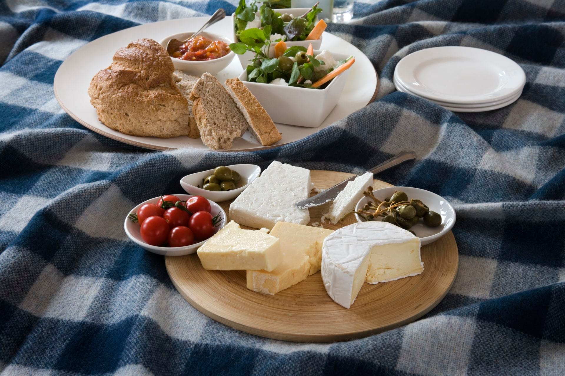 A rustic Welsh cheese board on a wooden platter over a blue and white checked wool blanket, featuring a wedge of crumbly white cheese, a block of pale cheddar-style cheese and a soft white-rinded round with a wedge cut away, alongside small bowls of cherry tomatoes, green olives and capers, with a plate of bara brith and sliced bread, chutney and salad visible in the background