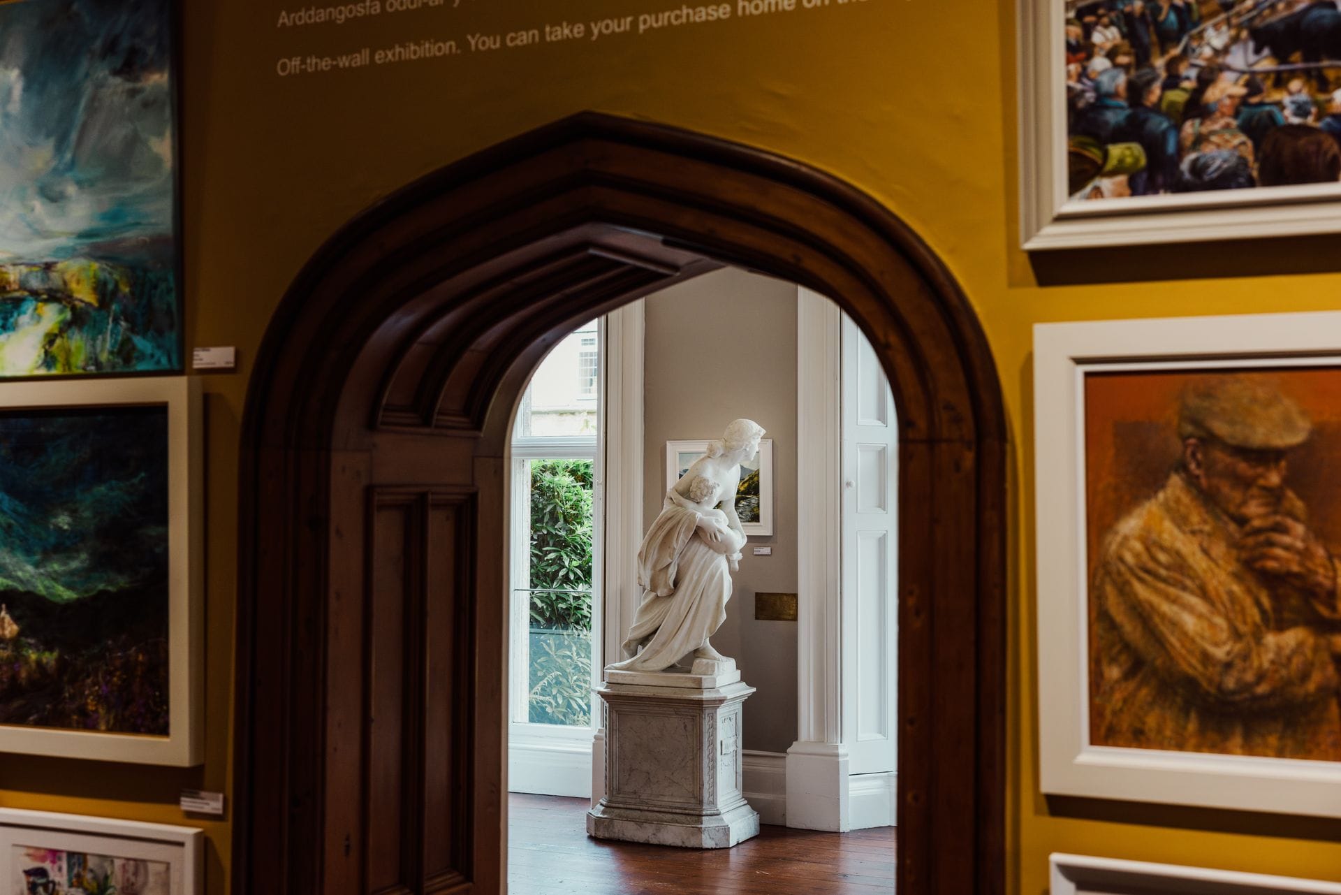 An interior view of the Plas Glyn art gallery in Wales, featuring mustard-yellow walls adorned with framed paintings and a prominent ornate dark wood archway looking through to a bright adjacent room displaying a classical white marble statue next to a window.