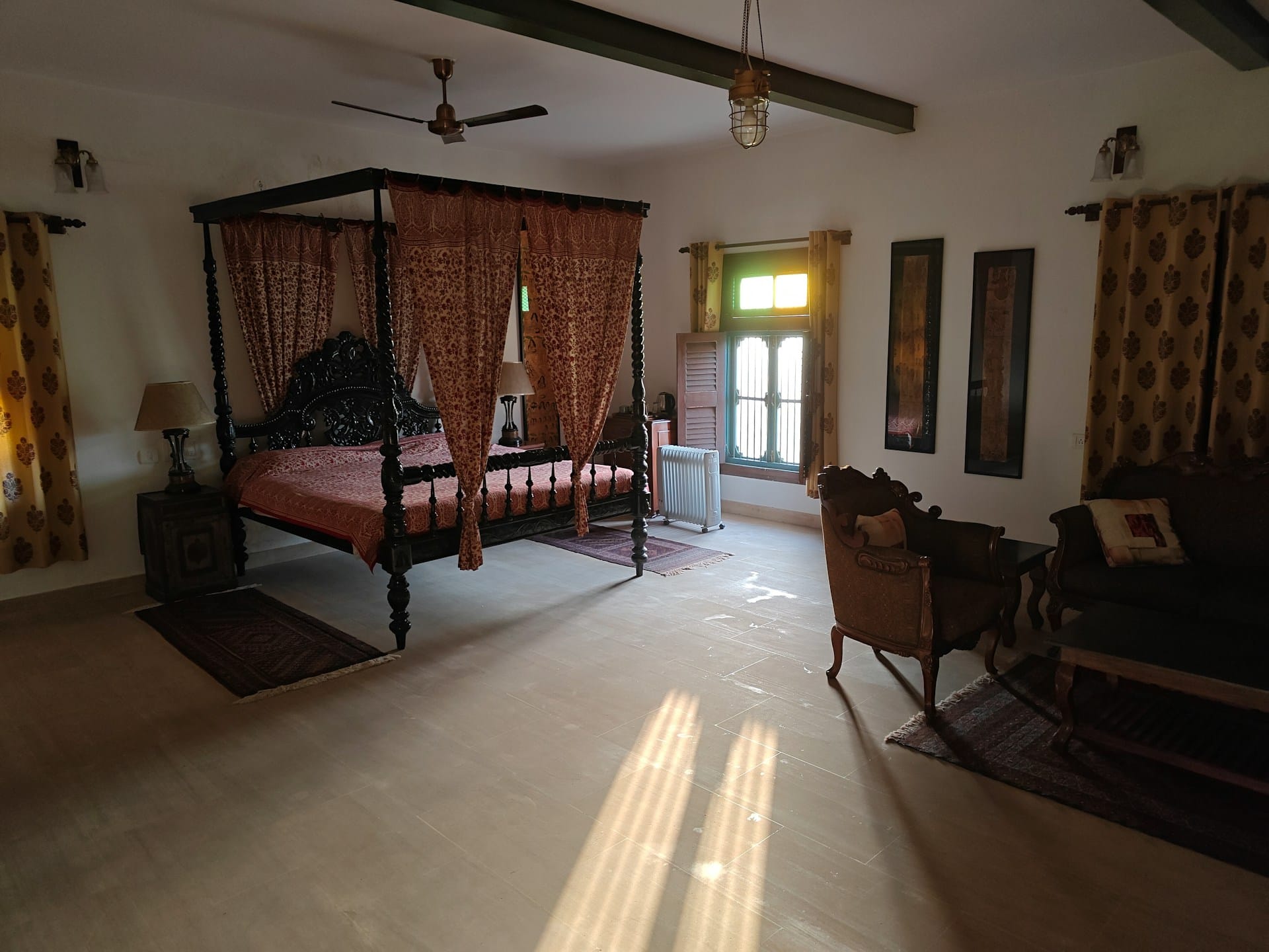 A spacious, luxury, rustic bedroom featuring a dark wood four-poster bed draped with patterned red curtains. Warm sunlight streams through a small window, casting long, bright rays across the light-coloured floor, highlighting a traditional seating area and dark wooden ceiling beams.