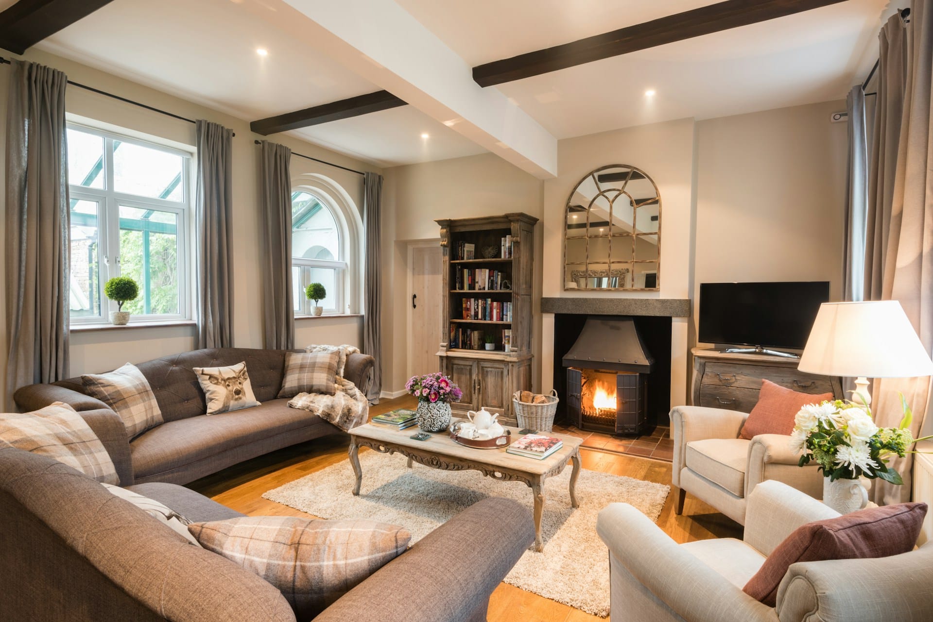 A cosy, well-lit luxury cottage living room featuring warm wooden floors and dark ceiling beams. A large grey corner sofa with plaid cushions and two light-coloured armchairs are arranged around a central coffee table and rug. In the background, a log burner glows warmly in the fireplace beneath a large decorative mirror, with natural light streaming in through large windows on the left.