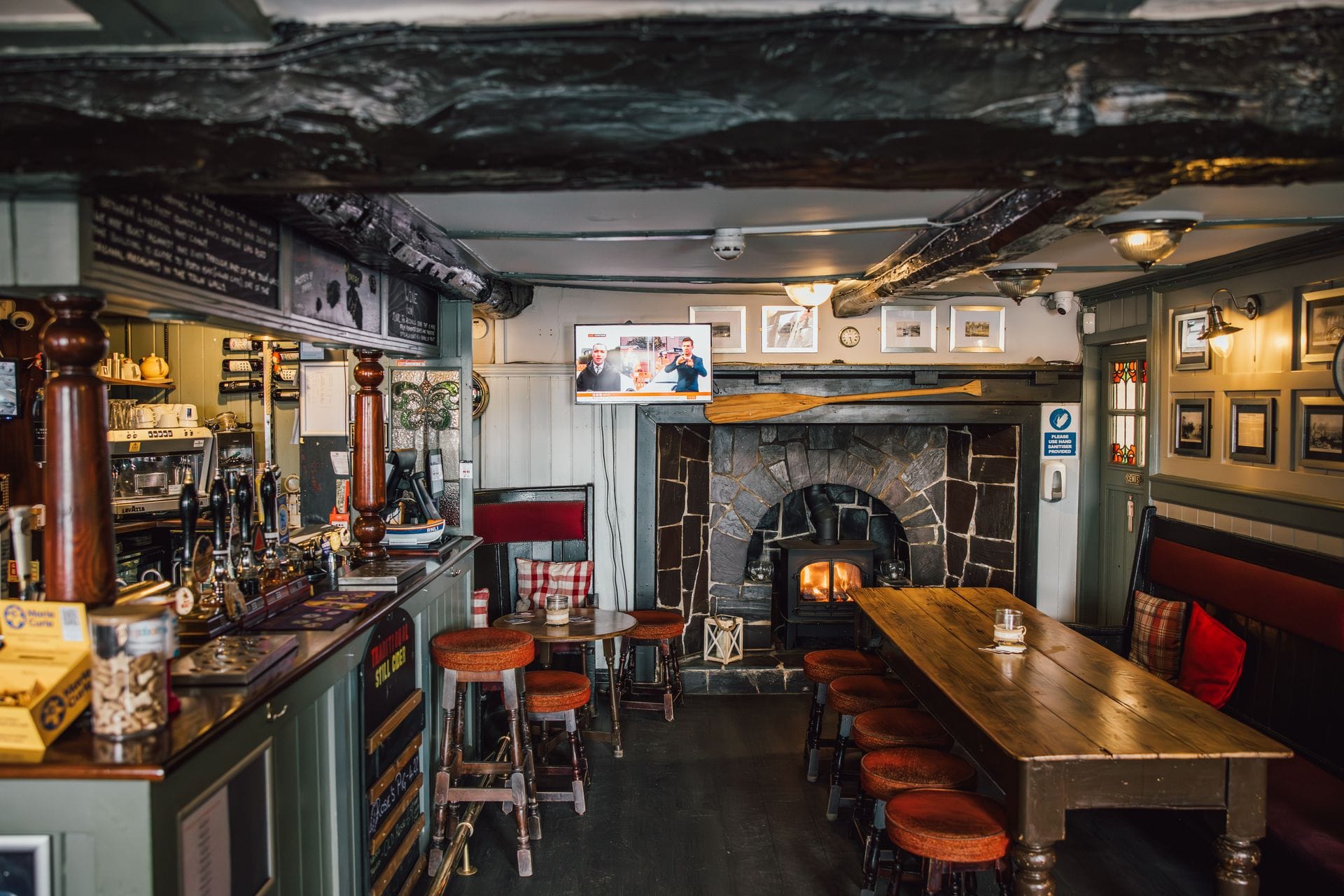 The warm, inviting interior of a traditional Welsh country pub. It features low, dark wooden ceiling beams, a large stone fireplace with a glowing wood-burning stove, a long communal wooden table with stools, and a classic bar area on the left.
