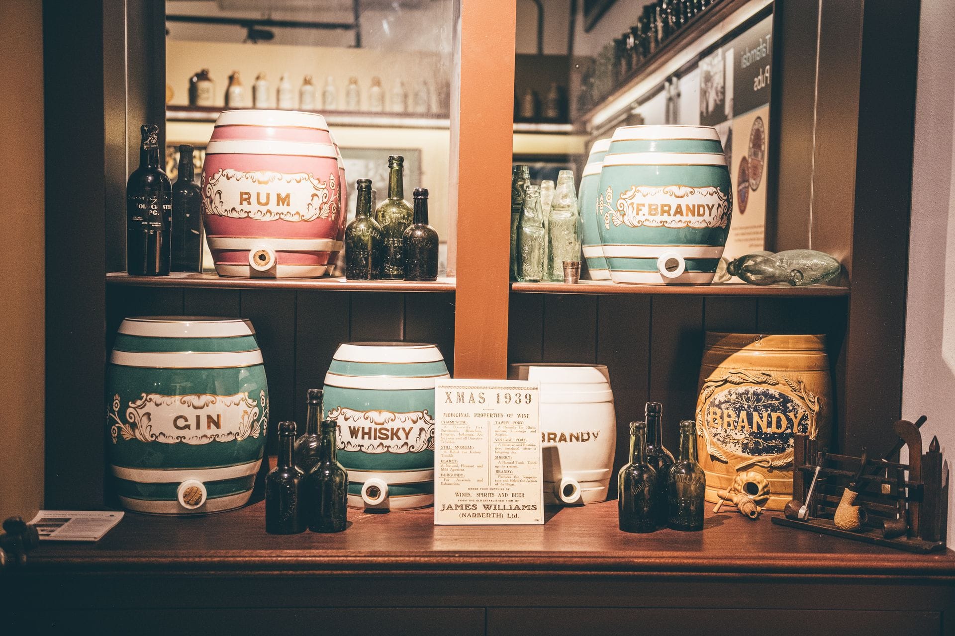 A historic display highlighting the rich heritage of Distilleries in Wales. A rustic wooden shelving unit holds a collection of vintage ceramic spirit dispensers, intricately painted in pastel colors with traditional labels for Rum, Gin, Whisky, and Brandy. Antique dark glass bottles and a small historical sign reading 'XMAS 1939' sit among the casks, showcasing the long-standing tradition of Welsh spirit making.