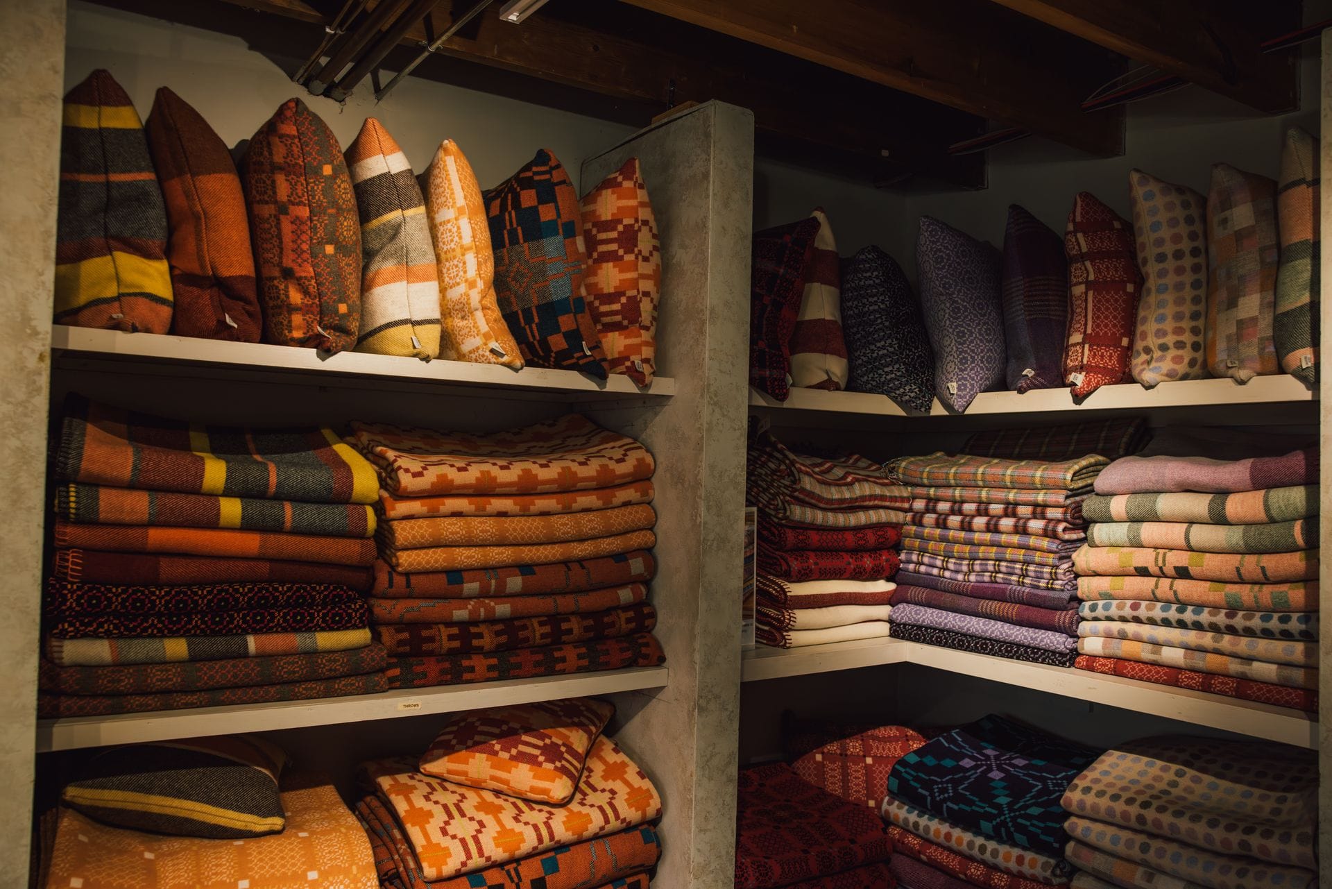 Shelves of folded Welsh wool blankets and cushions in traditional double-cloth tapestry patterns, in shades of orange, mustard, burgundy and purple, stacked in a mill shop.