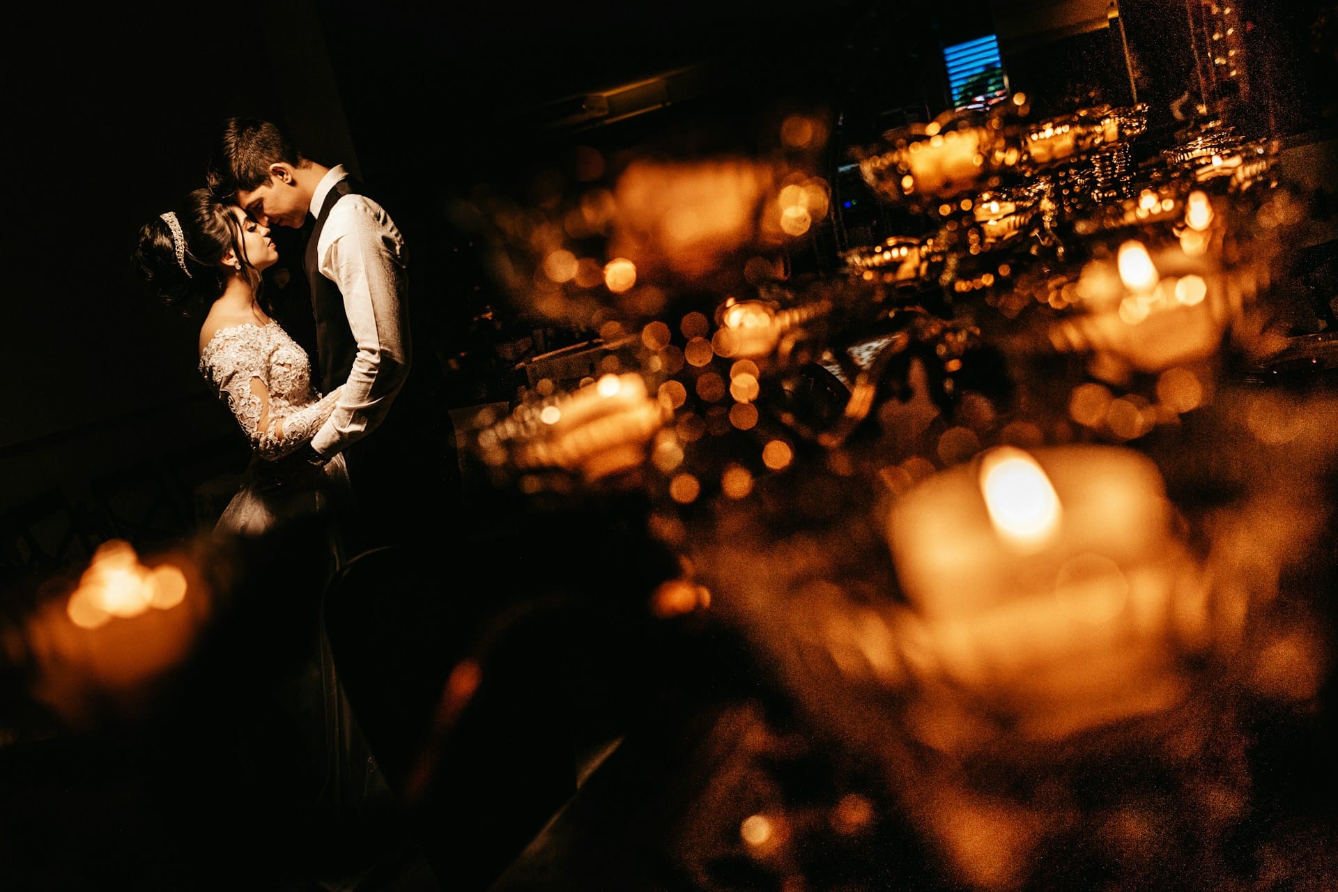 A wedding photographer capturing an intimate quiet moment, the couple pressed forehead-to-forehead in soft candlelight from a nearby wooden dining table set for a wedding reception, with warm golden light illuminating them — a documentary-style wedding photography moment.