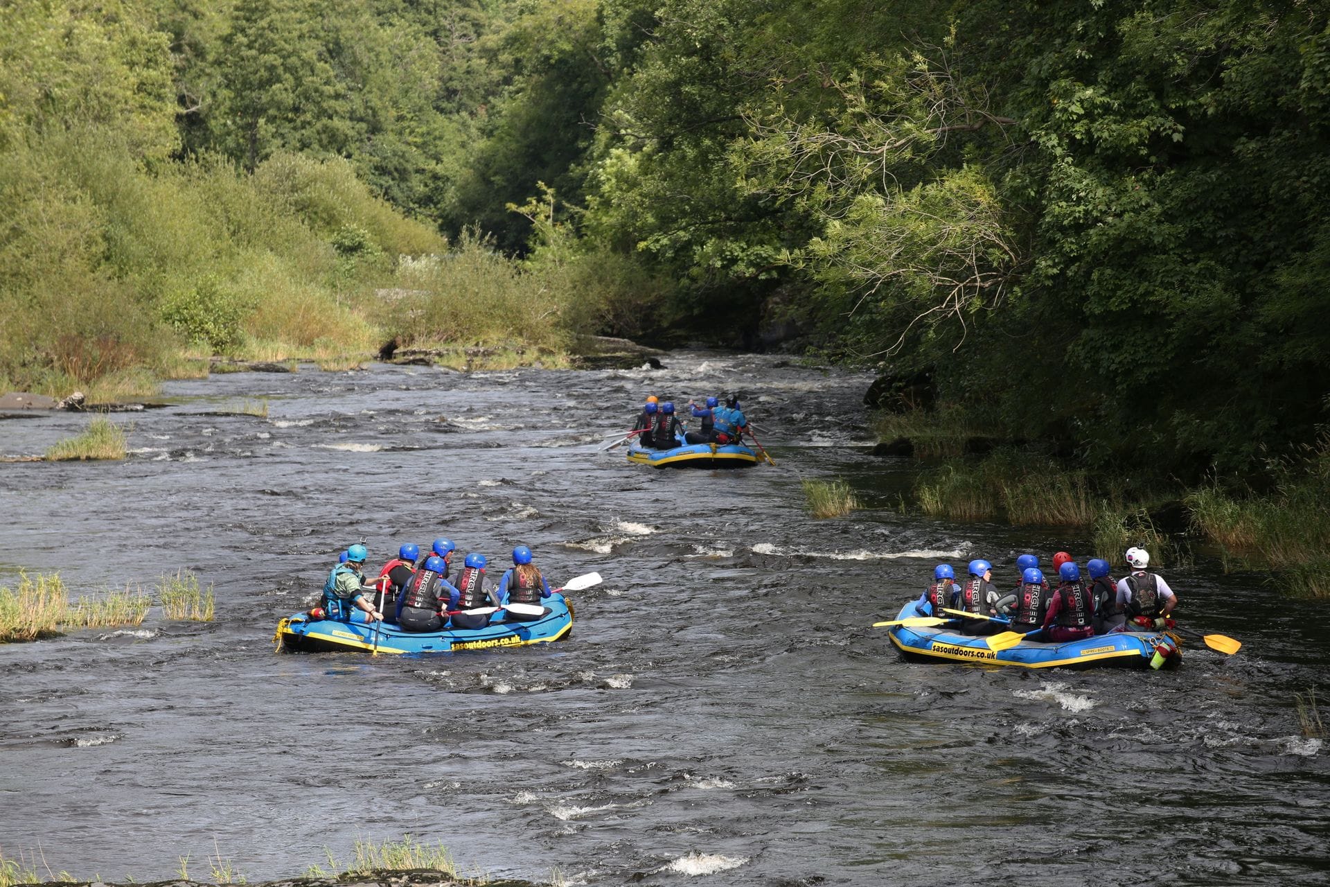 A scenic view of three blue and yellow inflatable rafts filled with adventurers wearing helmets and life jackets as they navigate rocky rapids. They are enjoying a thrilling white water rafting experience on the River Dee near Llangollen, surrounded by dense, lush green woodland along the riverbanks.