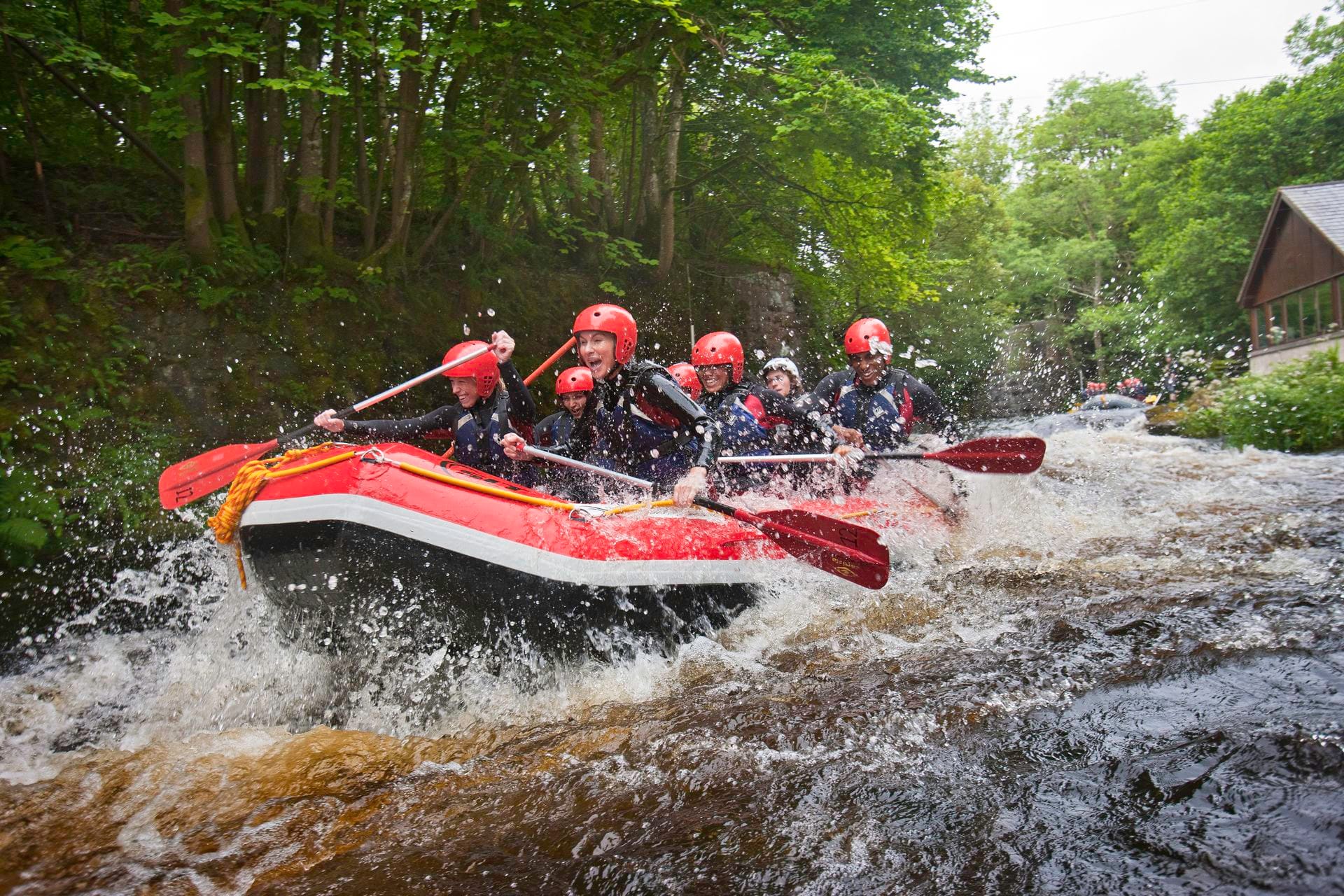 A team of adventurers wearing red helmets and wetsuits paddling a bright red raft through fast-moving, splashing rapids while white water rafting at Canolfan Tryweryn in Wales, surrounded by lush green woodland.