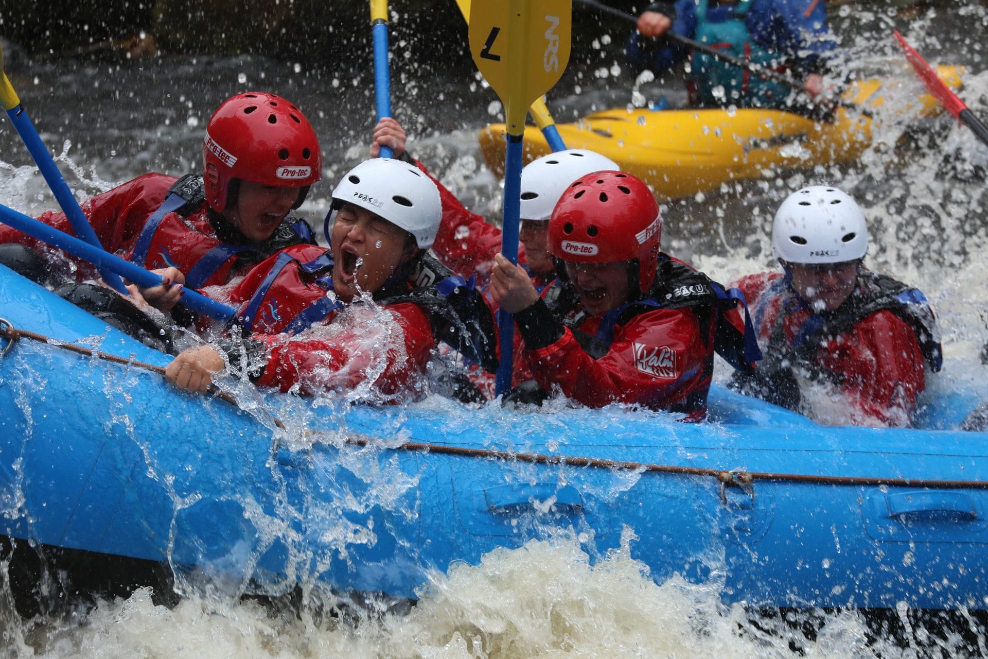 A thrilling close-up action shot of a group wearing red waterproofs and helmets, paddling a blue raft through crashing rapids while white water rafting at the National White Water Centre in Bala, Wales.