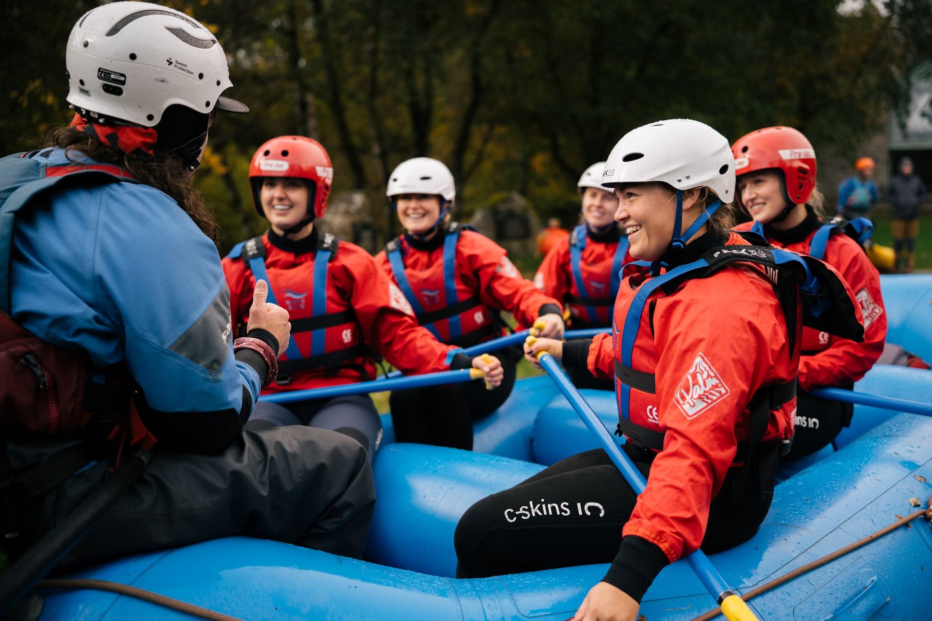 A cheerful group of people wearing red waterproof jackets and safety helmets sitting in a bright blue raft, holding paddles and smiling as they listen to their guide give instructions before setting off on a white water rafting adventure in Wales.