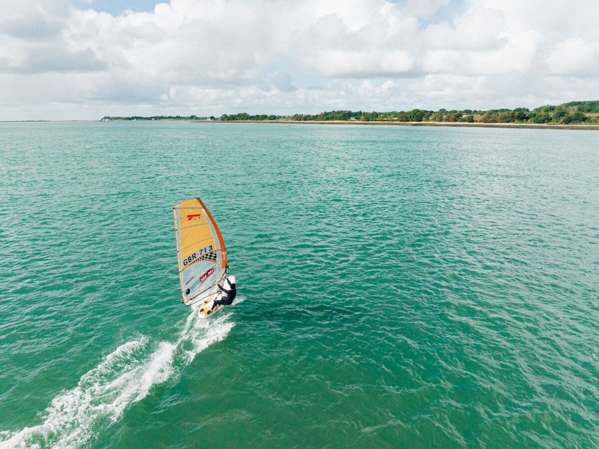 An aerial view of a person windsurfing on bright turquoise water in Wales, heading towards a lush green shoreline under a partly cloudy sky.
