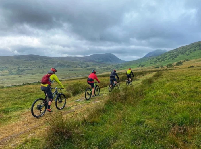 Four women mountain biking in Wales ride along a grassy trail through a wide, green valley, with hills and cloudy skies in the background.