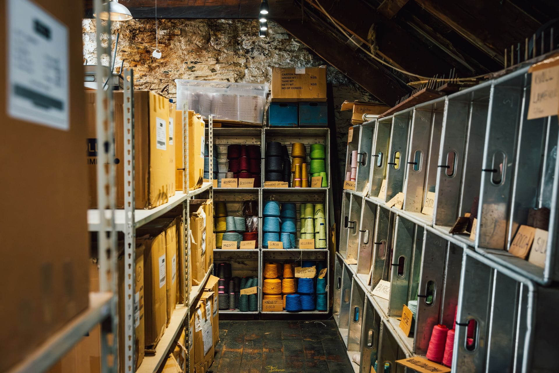 Rows of colour-coded wool yarn cones stored on metal shelving in a mill's loft store, labelled with shade names including Mojito, Sienna, Glacier and Harvest Gold, alongside stacked cardboard delivery boxes.