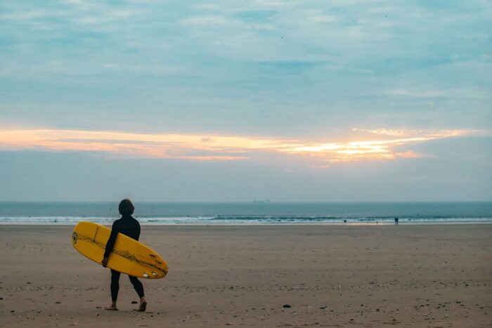 A person in a wetsuit stands on a sandy beach in Wales, holding a yellow surfboard and looking towards the sea under a cloudy sky at sunset, ready for surfing in Wales.
