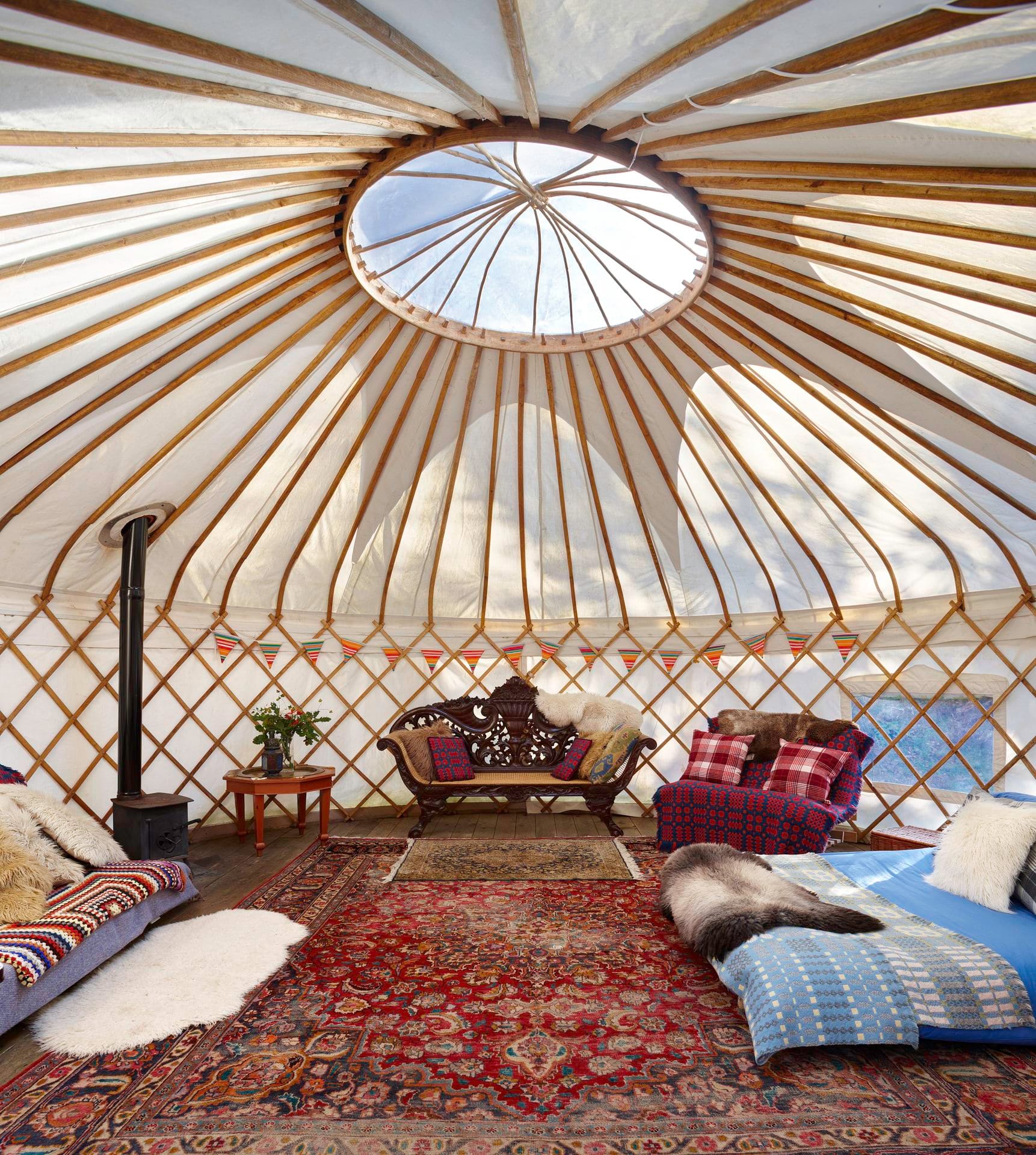 A bright, spacious interior view of a yurt. The structure features wooden lattice walls and roof poles radiating upwards to a central circular skylight. The wooden floor is heavily layered with a large, intricate red and blue Persian-style rug. The cozy room is furnished with a black wood-burning stove, an ornately carved dark wood bench, a red patterned armchair, and low beds or sofas adorned with colorful blankets, plaid cushions, and fluffy faux fur throws. A string of colorful bunting decorates the lower lattice wall, and natural light casts gentle tree silhouettes on the white canvas roof.