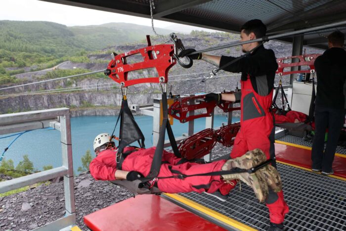 A person in red gear and a helmet is harnessed to a zip wire, preparing to launch over a blue lake, while another individual in red overalls assists—a true taste of adventure in Wales on a thrilling metal platform.