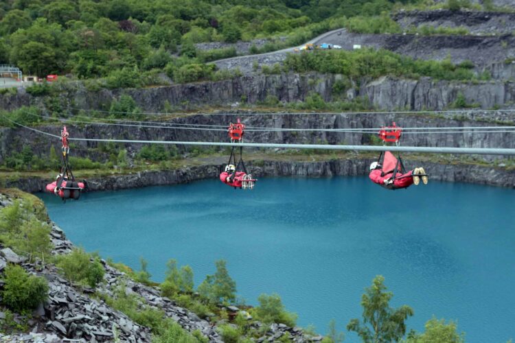 Three people in red suits and helmets ride zip wires over a large blue quarry lake, surrounded by rocky cliffs and green foliage