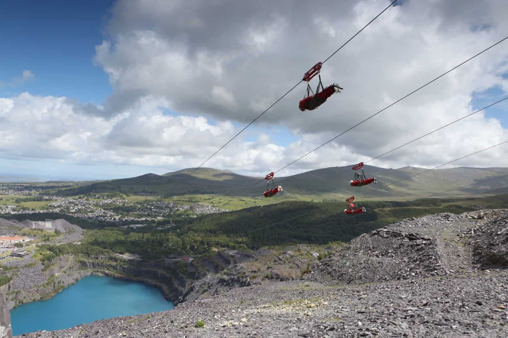 4 people going down a zip wire at zip world with the mountains in the background and a blue lake in the foreground underneath the riders